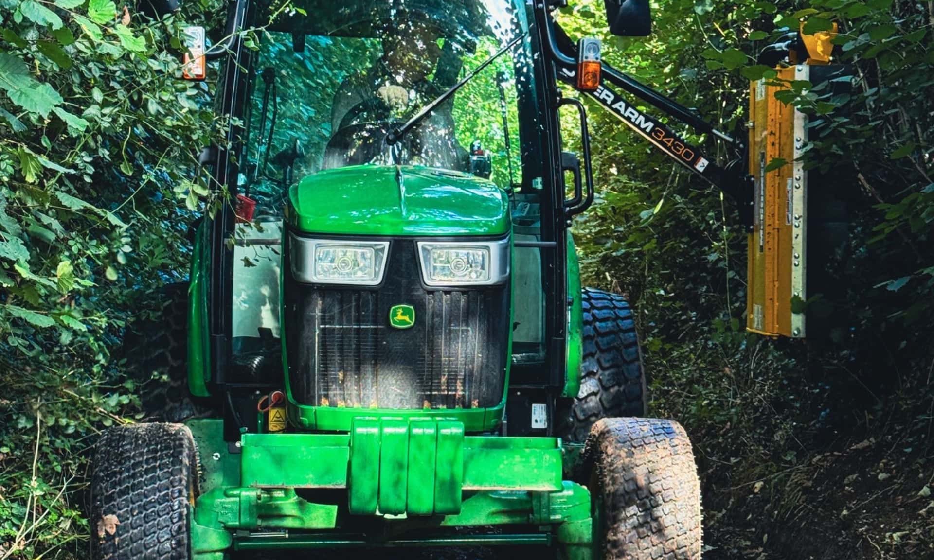 John Deere compact tractor with a McConnel PA3430 reach-arm flail cutting a hedge in a narrow Hampshire lane