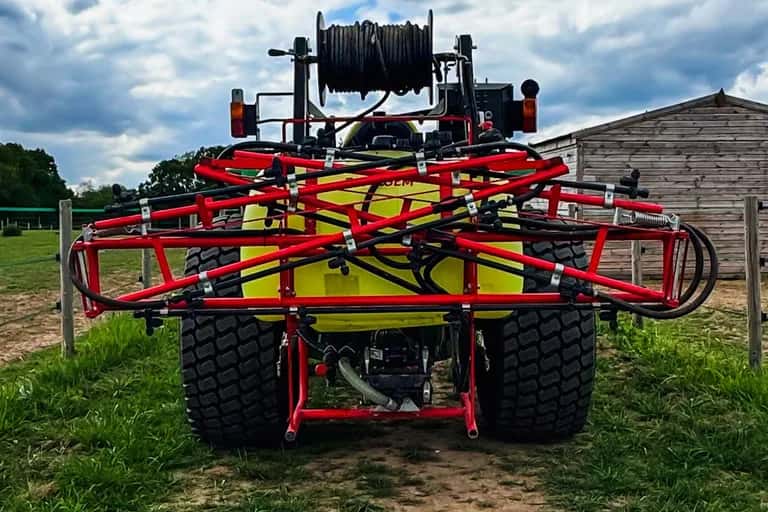 Rear view of a tractor-mounted boom sprayer with its booms folded, parked beside a paddock fence under a cloudy sky