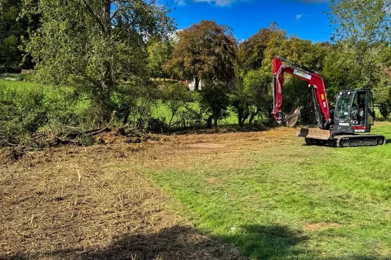 Excavator working across a cleared East Meon paddock strewn with chipped brash and excavated root material